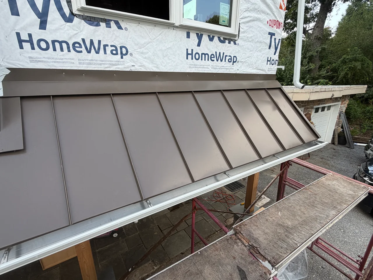 Brown standing-seam metal roof with a silver gutter beneath a window and exposed white house wrap, with wooden posts and red scaffolding with worn plywood platforms below